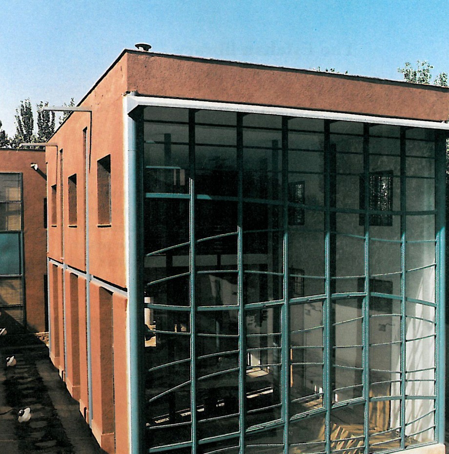 Three-storey brick villa with a tall glass hall on the right wing, the glass animated by a curving blue-painted steel grid; ground floor exposed on columns over the pool.