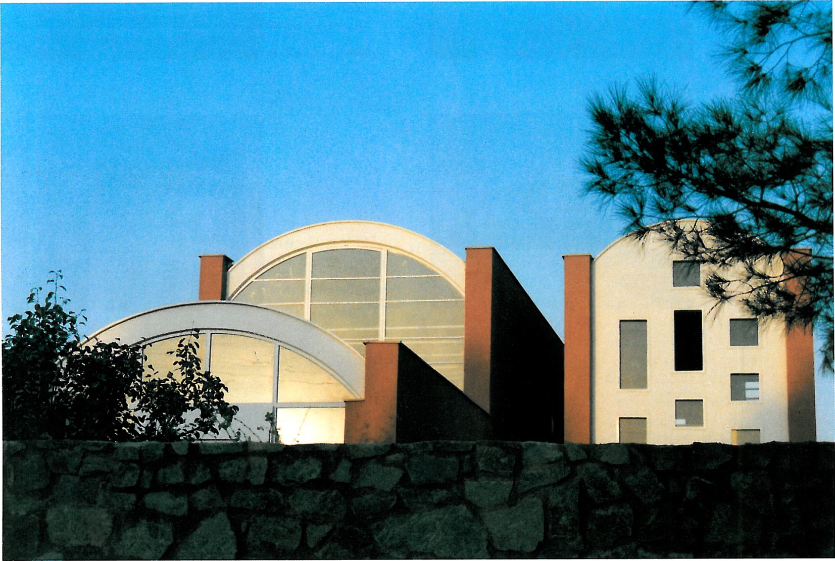 Two-volume residential building: a barrel-vaulted curved-roof common volume at left and a perforated brick private volume at right, separated by a narrow alley, in golden afternoon light.