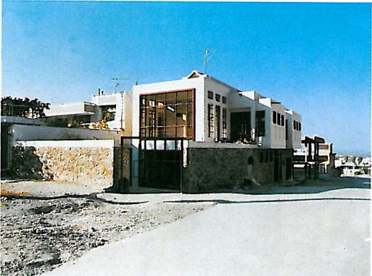 Low rectilinear white building with deeply shaded portico, standing alone on a sandy lot under a clear sky.