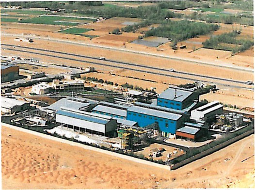 Single-storey industrial complex with white metal cladding and a red banded roof line, in arid open landscape.