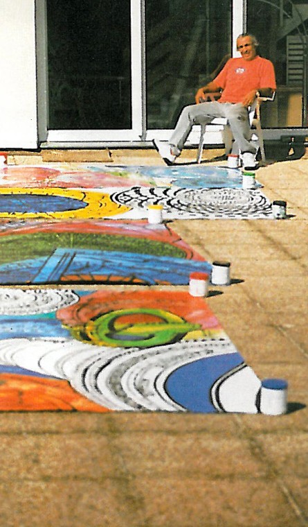 Black-and-white photograph of Charles-Hossein Zenderoudi seated on a giant painted-pencil sculpture, with canvas works spread on the ground in front of his studio.