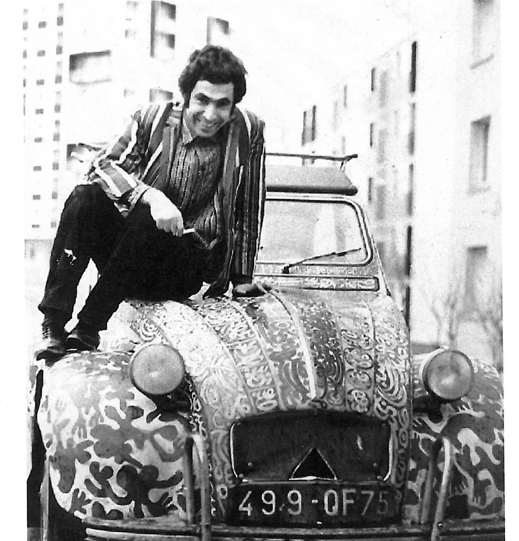 Photograph of Charles-Hossein Zenderoudi seated atop a Citroën 2CV whose body is entirely covered in his black-and-white calligraphic painting, French license plate visible.
