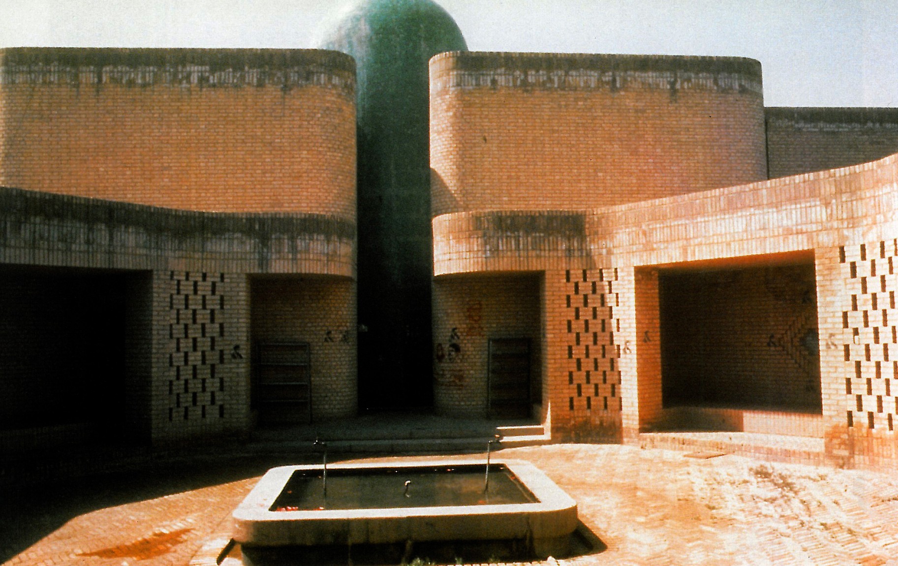 Photograph of the inner courtyard of the Jondi-shapur University Mosque with a stepped rectangular fountain, perforated brick walls and a green-domed minaret rising behind