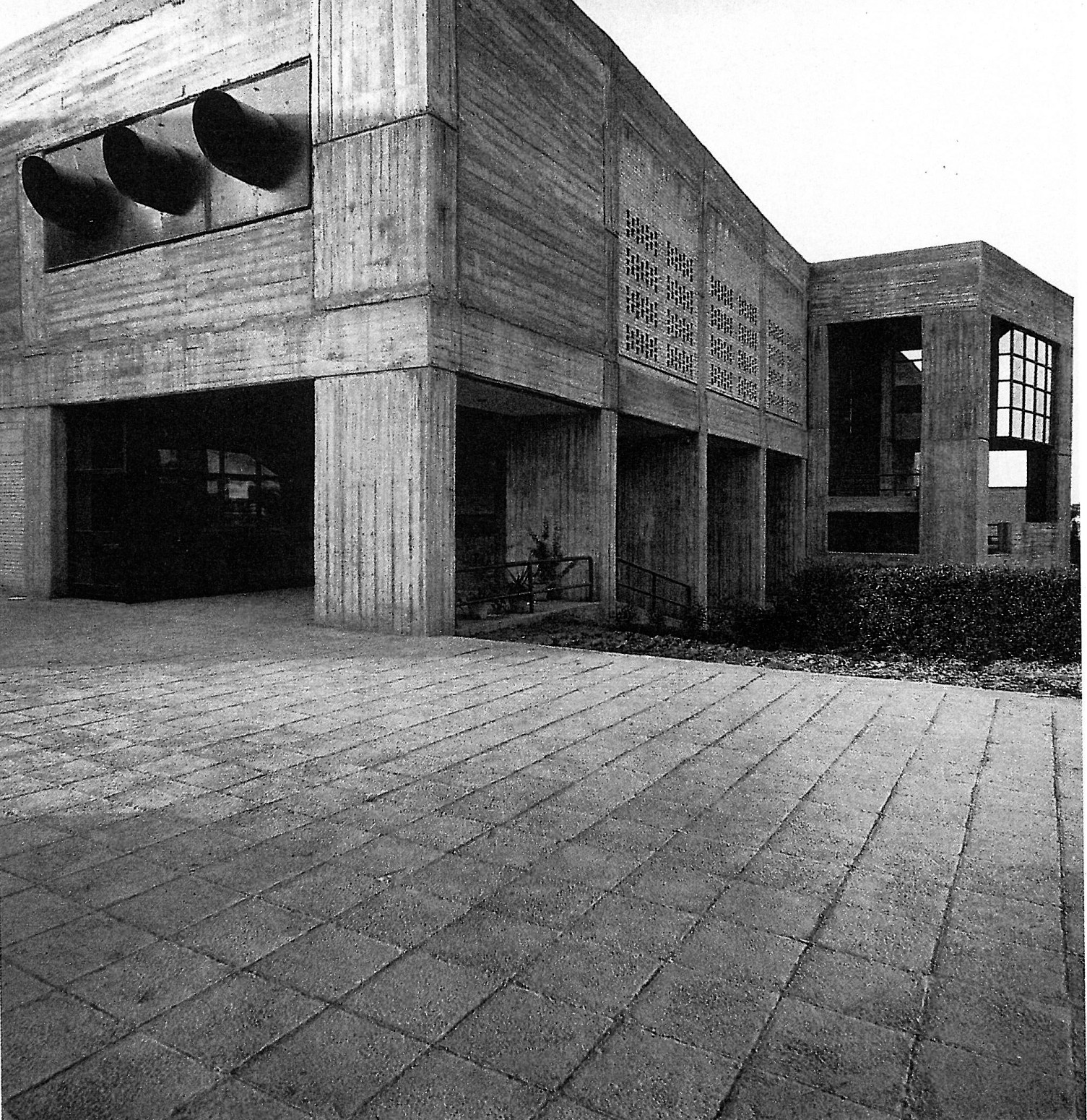 Daytime photograph of the Jondi-shapur University Gymnasium showing the brutalist concrete facade with deep circular openings on the upper level and exposed concrete frame at ground level