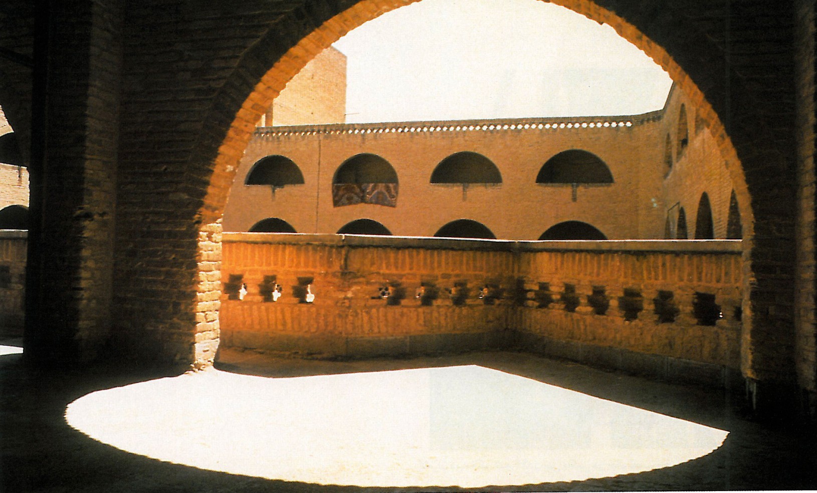 Interior photograph from beneath the brick vaulted gallery of a Shushtar New Town apartment building, showing the rhythm of arched openings looking out toward the neighborhood plaza