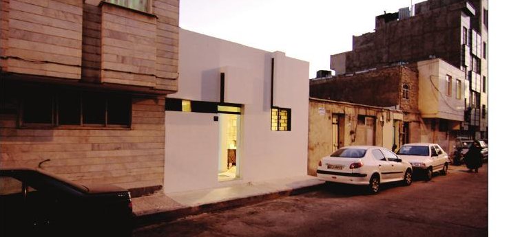 Wide dusk view of the alley with parked cars showing the white volume sitting between worn neighbouring houses