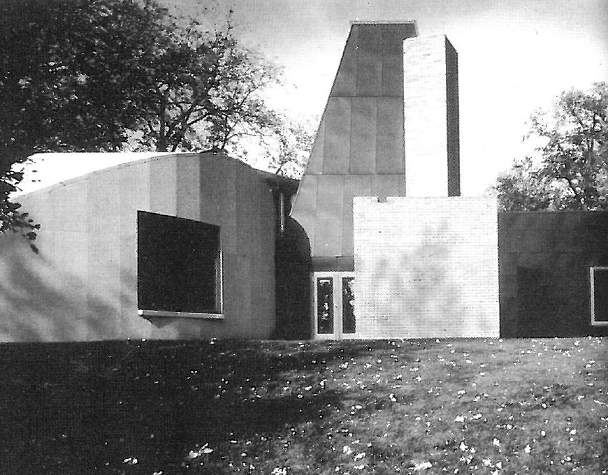 Photograph of a small Hejduk-designed chapel or house with sloped gable roof and dark cladding