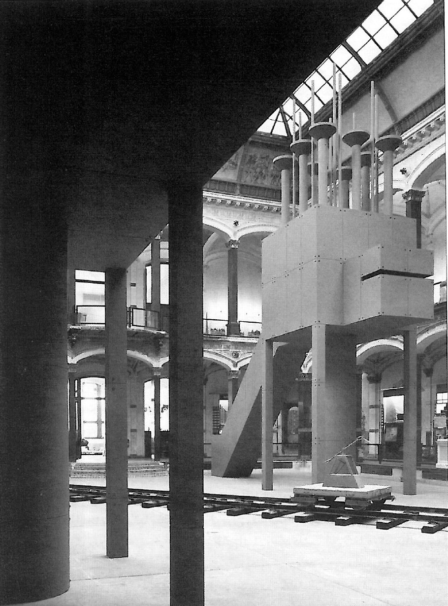 Interior installation photograph inside a brick-walled gallery with iron columns and freestanding architectural pieces