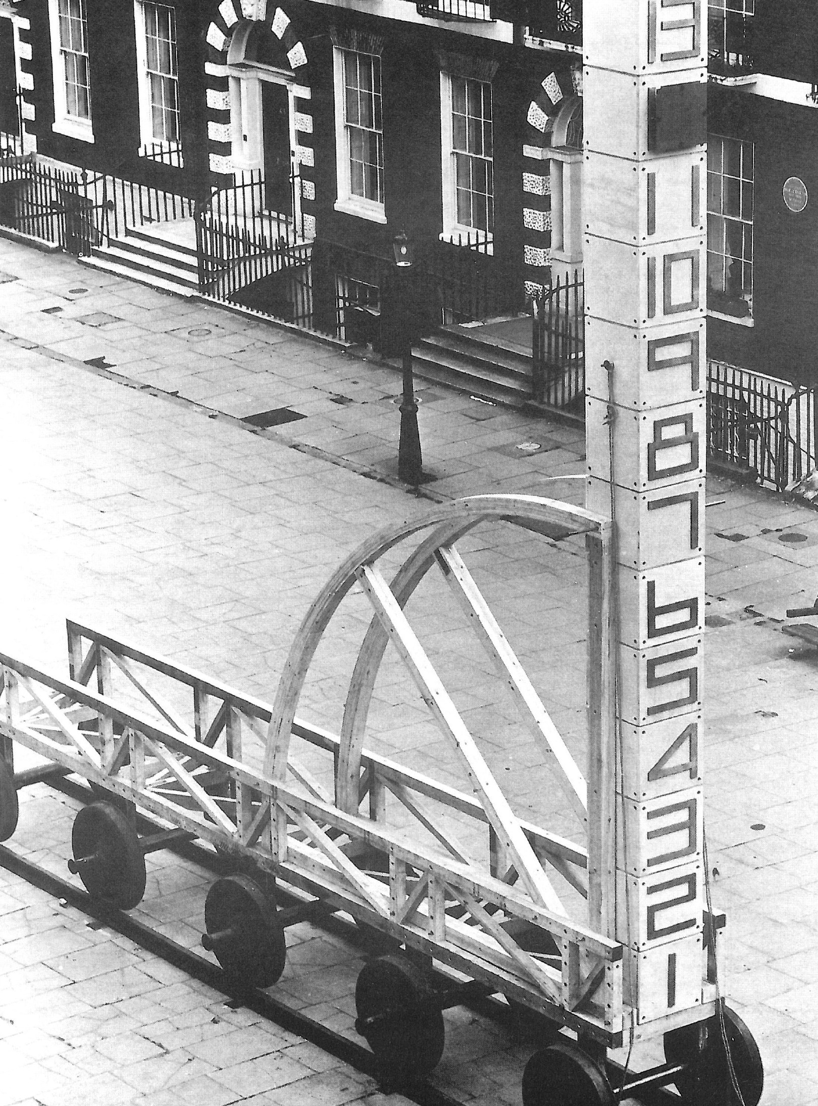 Full-page black-and-white photograph of Hejduk's vertical numbered architectural piece on wheels, parked on cobblestones in front of Georgian row houses