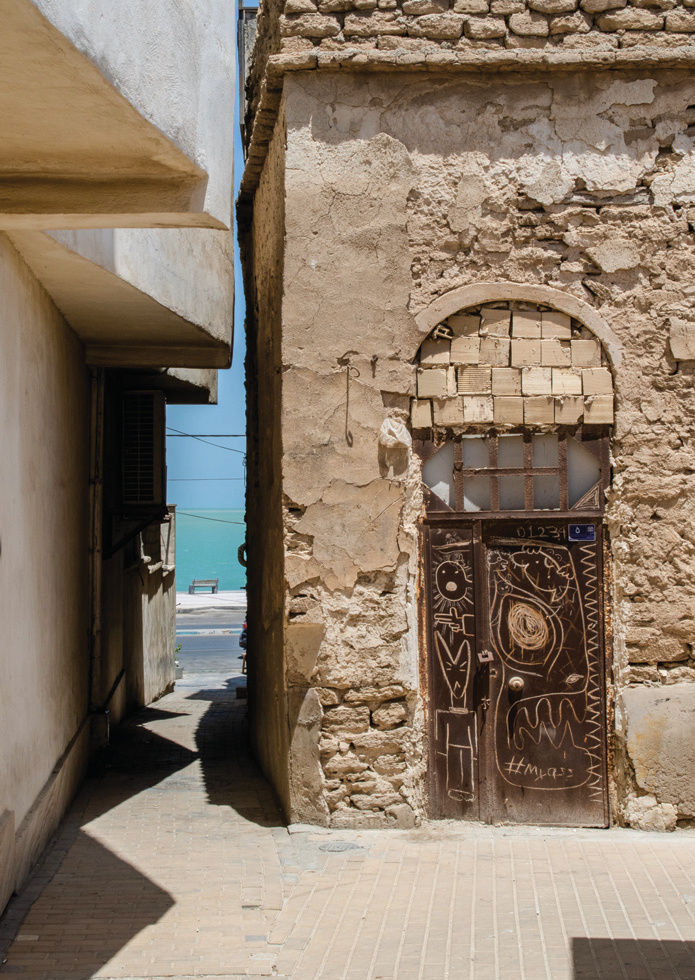An alley leading to the sea, with a graffiti-covered door and coral stone walls