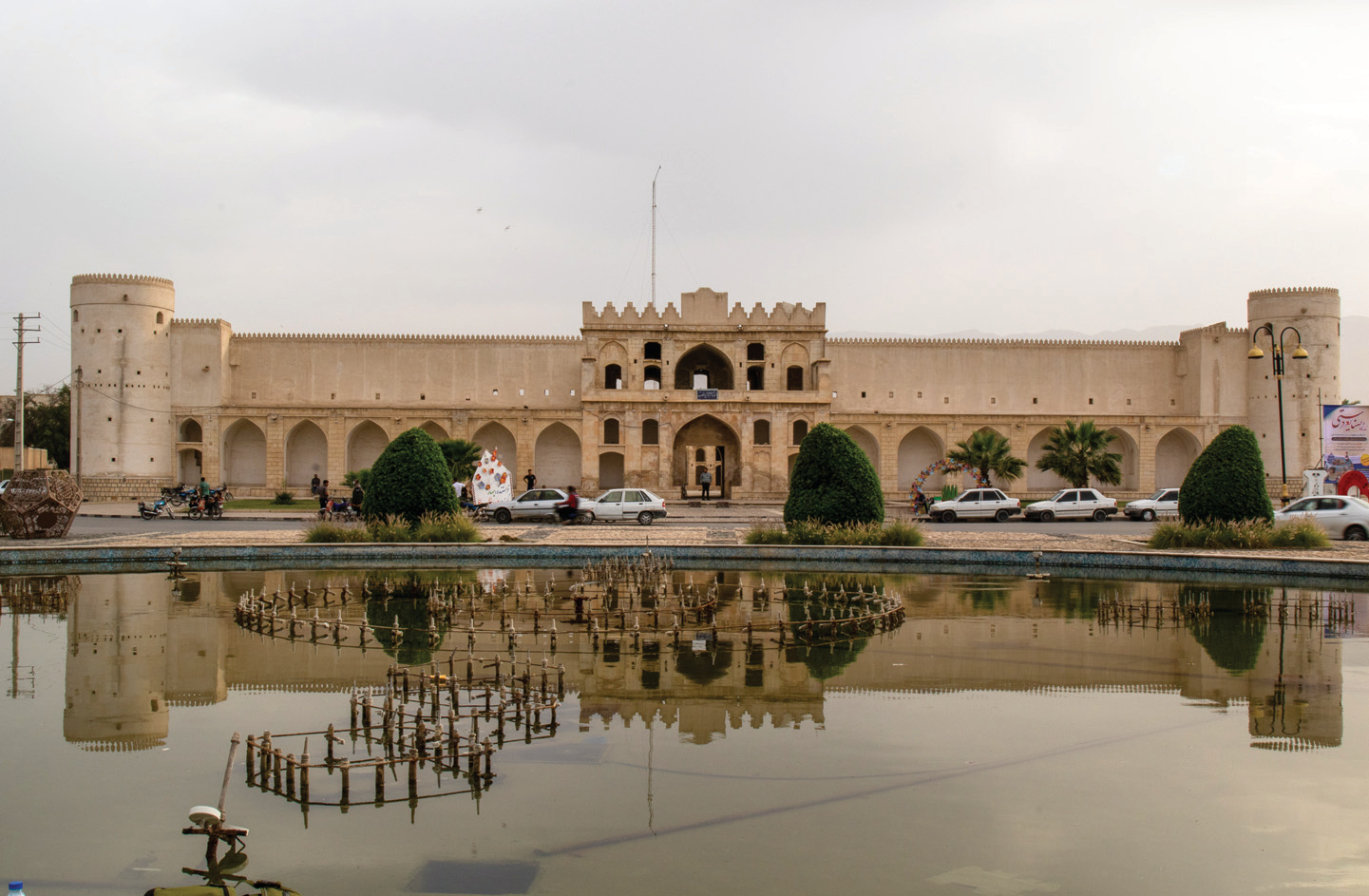Full exterior view of the Borazjan Caravanserai with corner towers, battlements, arched facade, trees, and reflecting pool in front