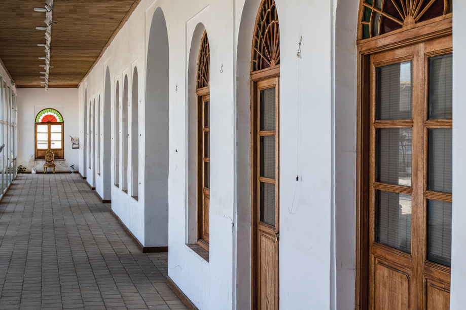 Long gallery corridor with arched recesses, wooden doors with fan lunettes, and a stained glass window at the end