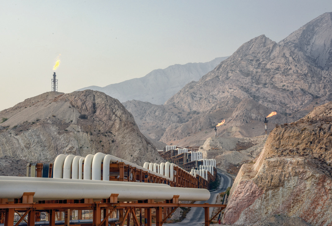 Gas pipelines threading through a mountain pass, with flares burning in the distance
