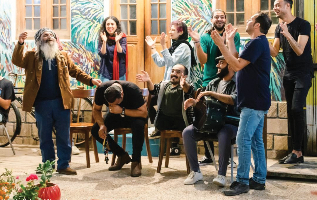 Musicians performing and an elderly bearded man dancing joyfully while young people clap at a Khayyami gathering