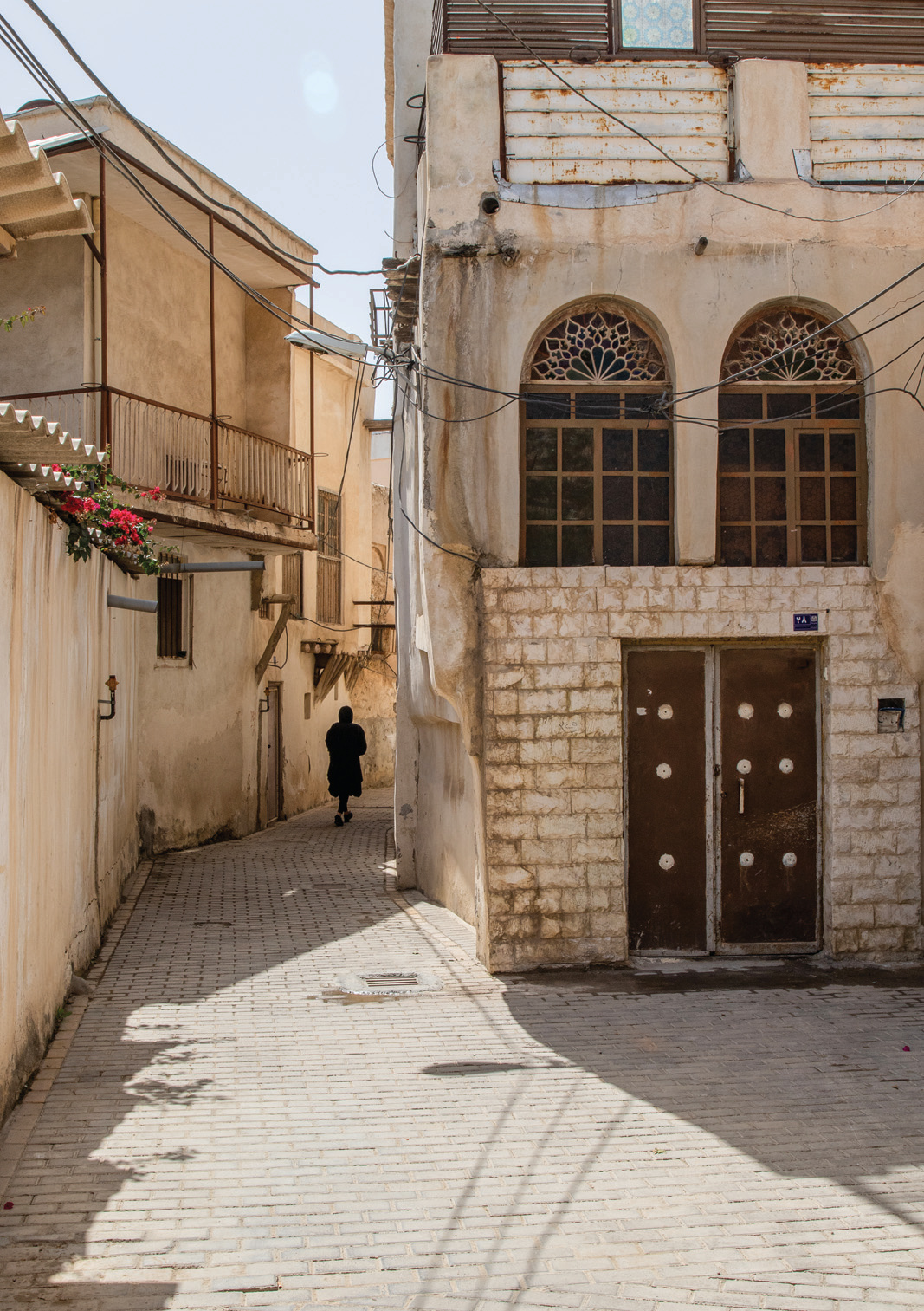A solitary figure walks beneath lattice-arched windows in a sunlit Bushehr alley