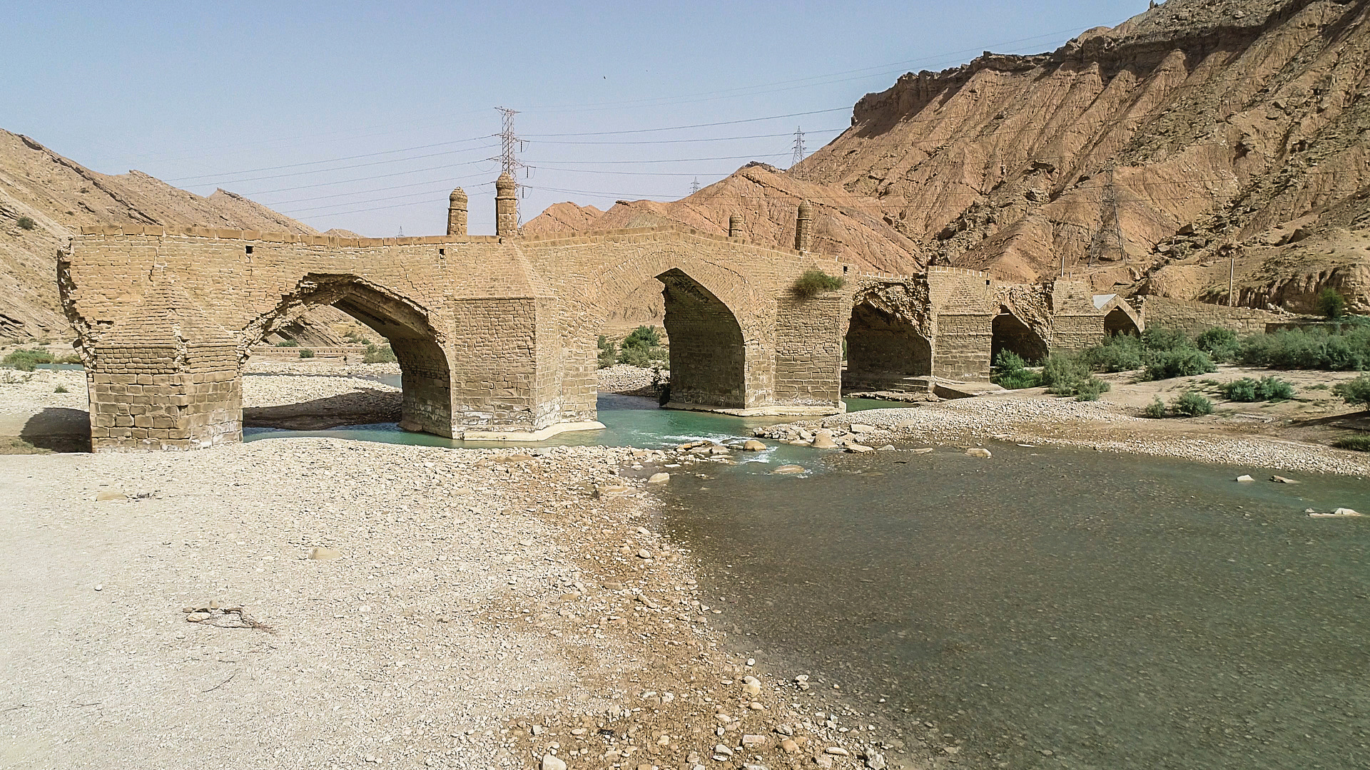 Moshir Bridge spanning the Dalaki River with six stone arches and turquoise water flowing beneath, red mountains behind