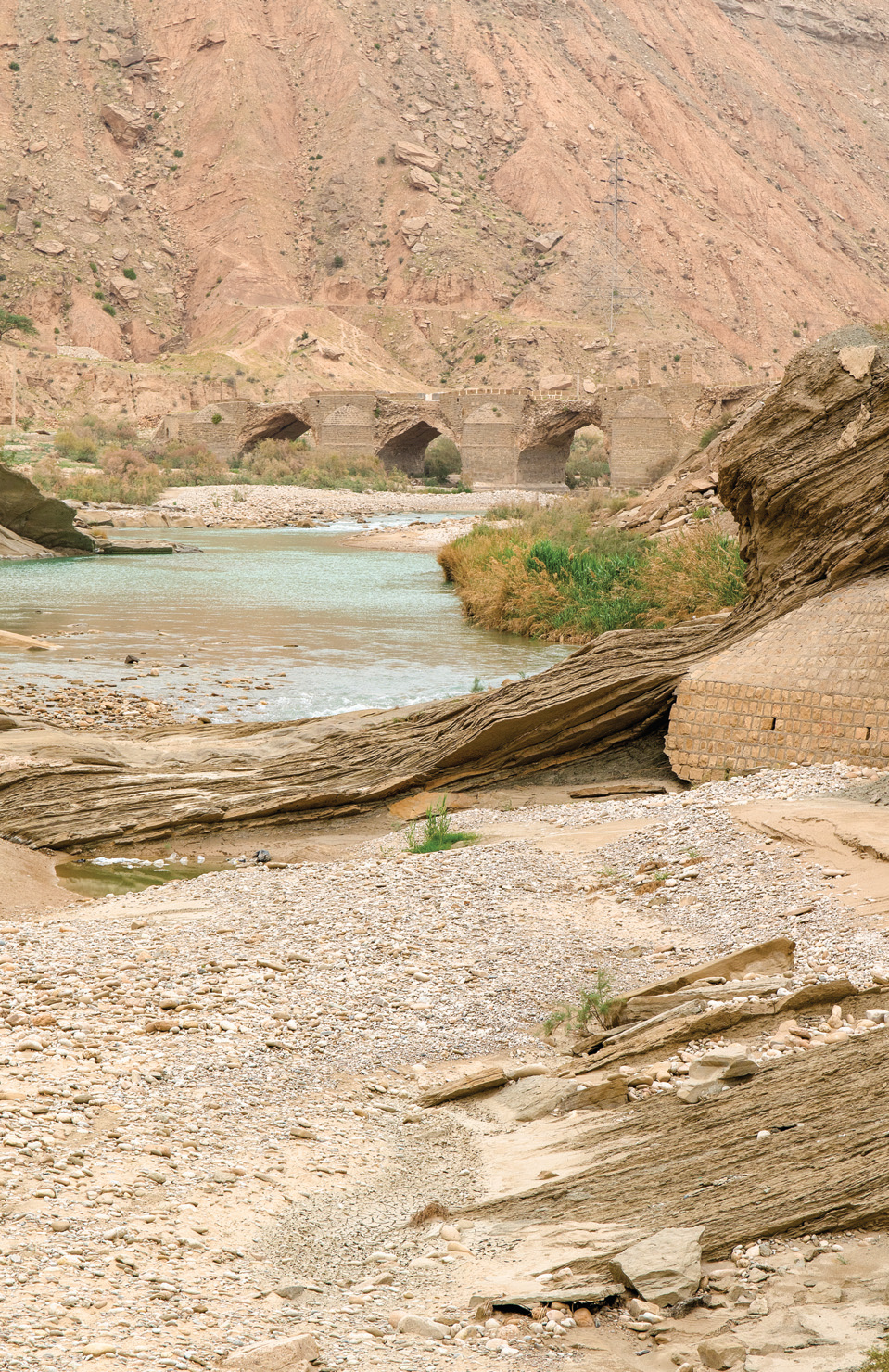 View along the Dalaki River toward the Moshir Bridge, with layered sedimentary rock formations in the foreground