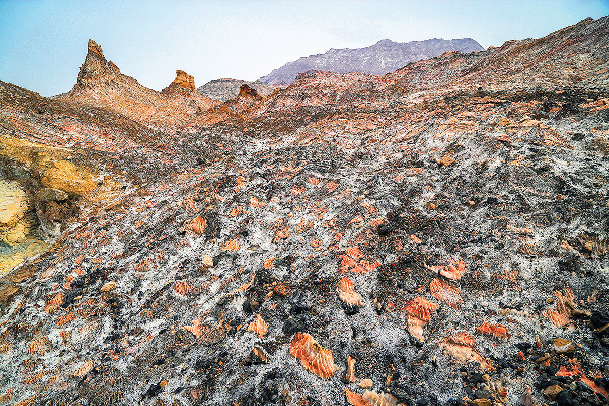 Panoramic view of the salt dome landscape with dark rock and vivid orange mineral deposits, salt-covered peaks in background