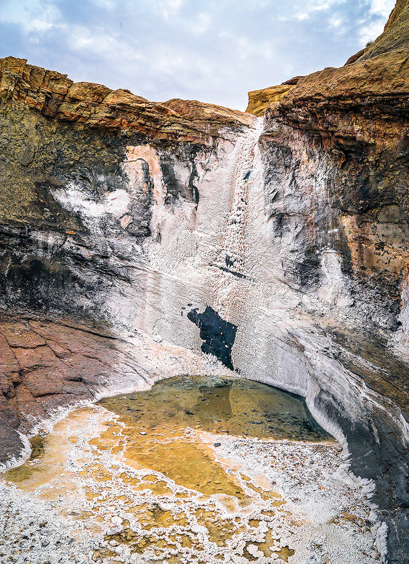 A salt waterfall flowing down a cliff face into a golden-brown pool below, surrounded by layered rock walls