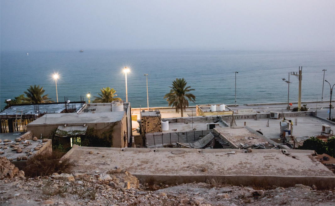 View at dusk over Siraf's flat rooftops toward the Persian Gulf, with streetlights and palm trees