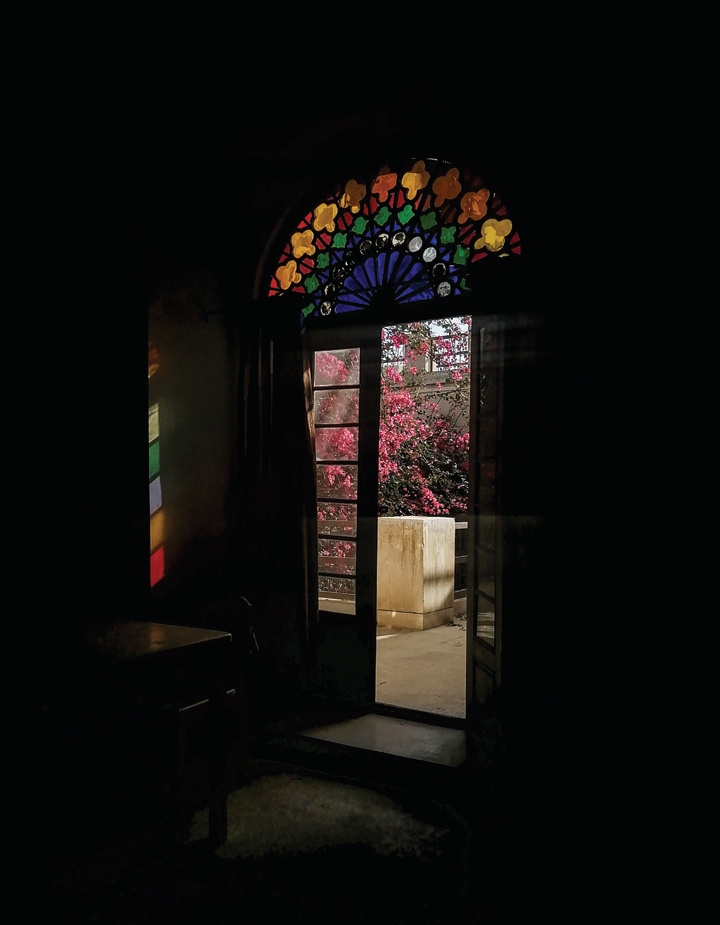 View from a dark interior through an open wooden door with a colorful stained glass lunette, looking out to the courtyard and bougainvillea