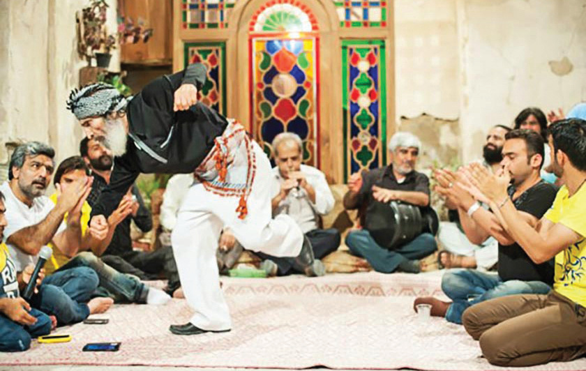 An elderly man performing a traditional dance in a historic Bushehri room with stained glass windows, while seated men clap rhythmically
