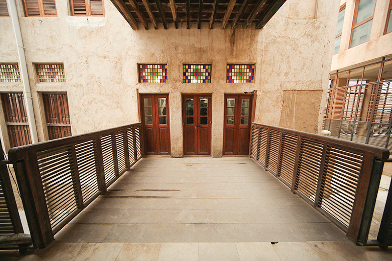 Upper floor gallery with three wooden doors, colorful stained glass lunettes above, and wooden louvered railing