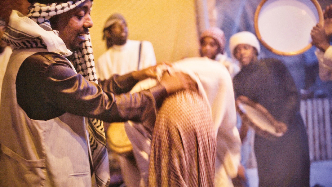 A man guiding a covered participant during the Zar ceremony while musicians play drums in the background