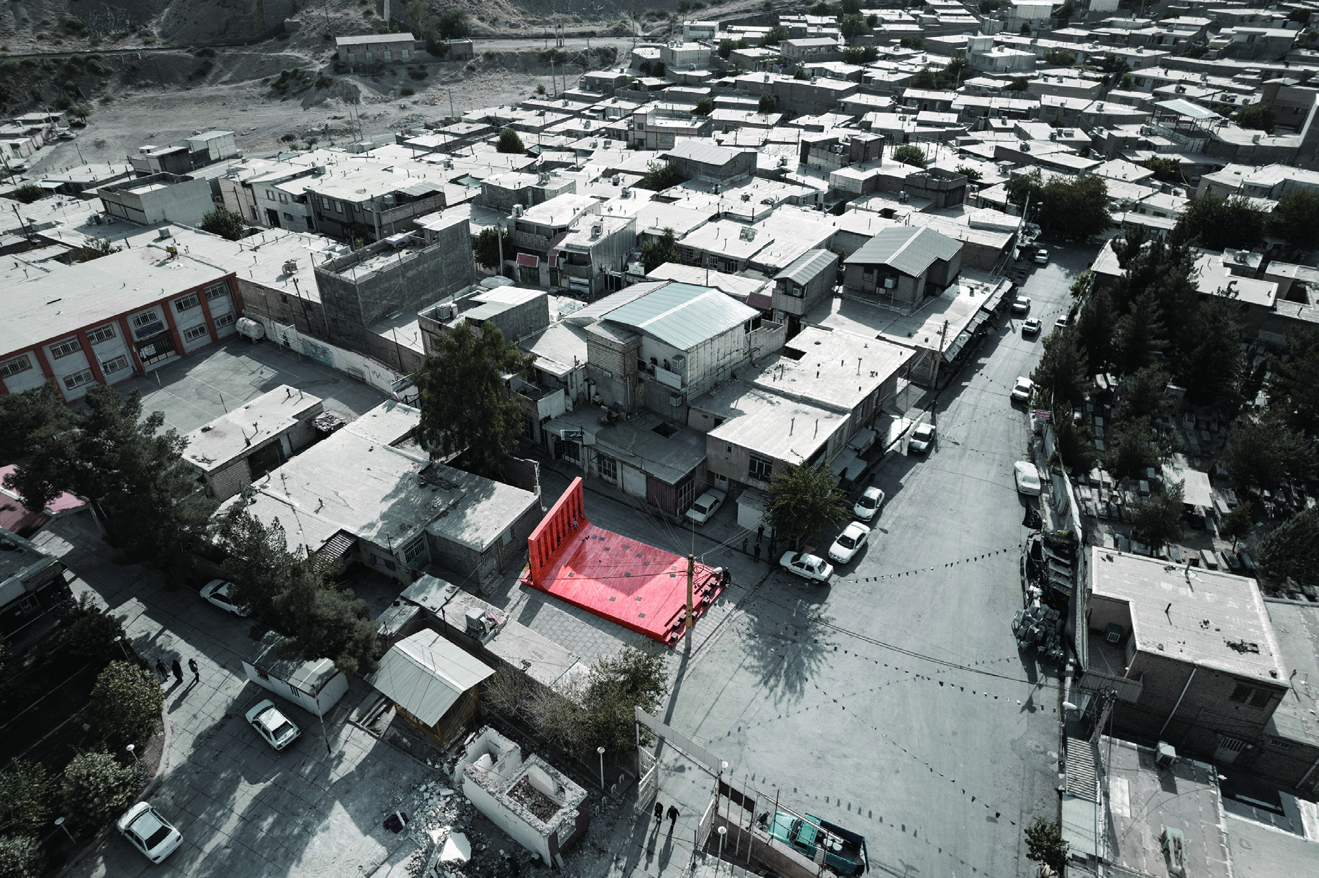 Aerial view of Sepiddasht with the red stone platform highlighted in color against the desaturated surrounding town fabric, showing how the project stands out in the urban landscape