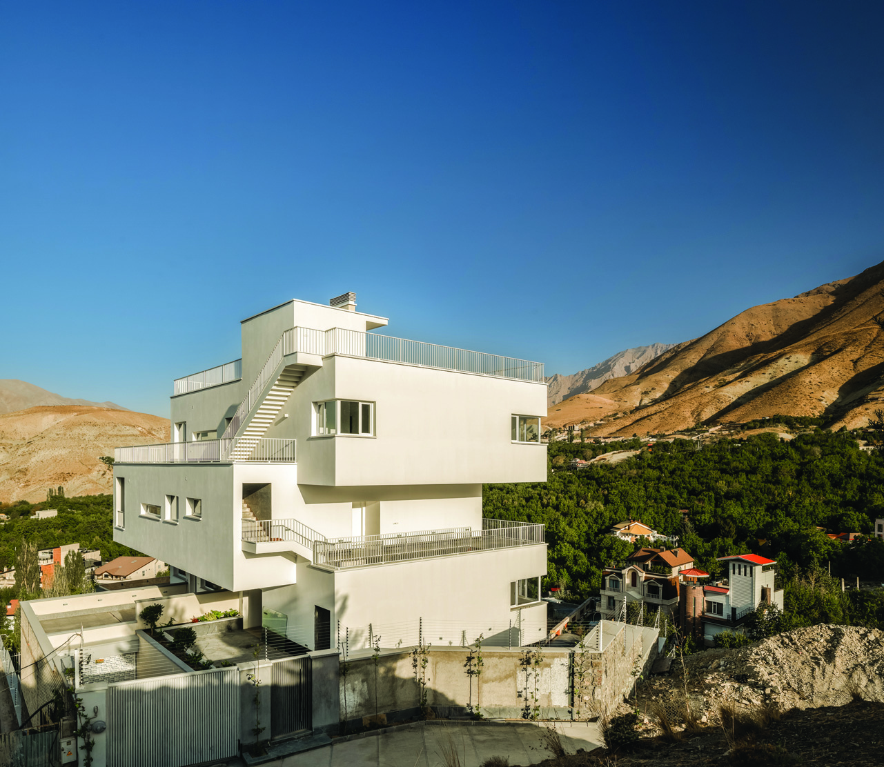 The building seen from a distance on the hillside of Lavasan, showing it perched above an exposed rock cliff with the white volumes stacking upward