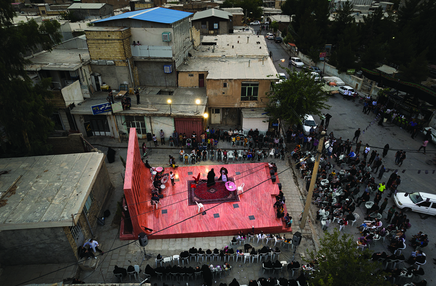 Aerial view of a cultural performance at dusk on the red platform, surrounded by the townspeople sitting in chairs and standing along the edges, with the town's buildings and rooftops serving as additional vantage points