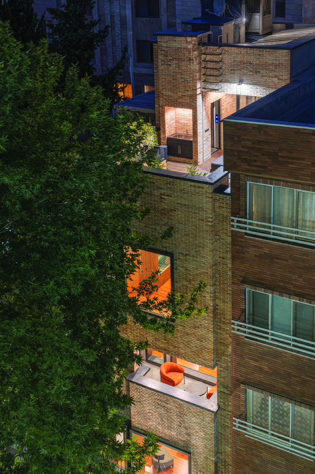 Aerial night view of House No. 10 showing the brick facade, illuminated terraces at multiple levels, an orange armchair on one terrace, rooftop pool area, and a large tree alongside the building