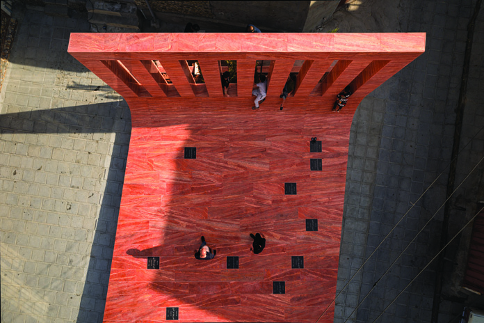 Overhead aerial view of the red stone platform showing the vertical wall at the top with openings, the curving transition to the ground plane, black grave markers embedded in the surface, and a person sitting on the platform