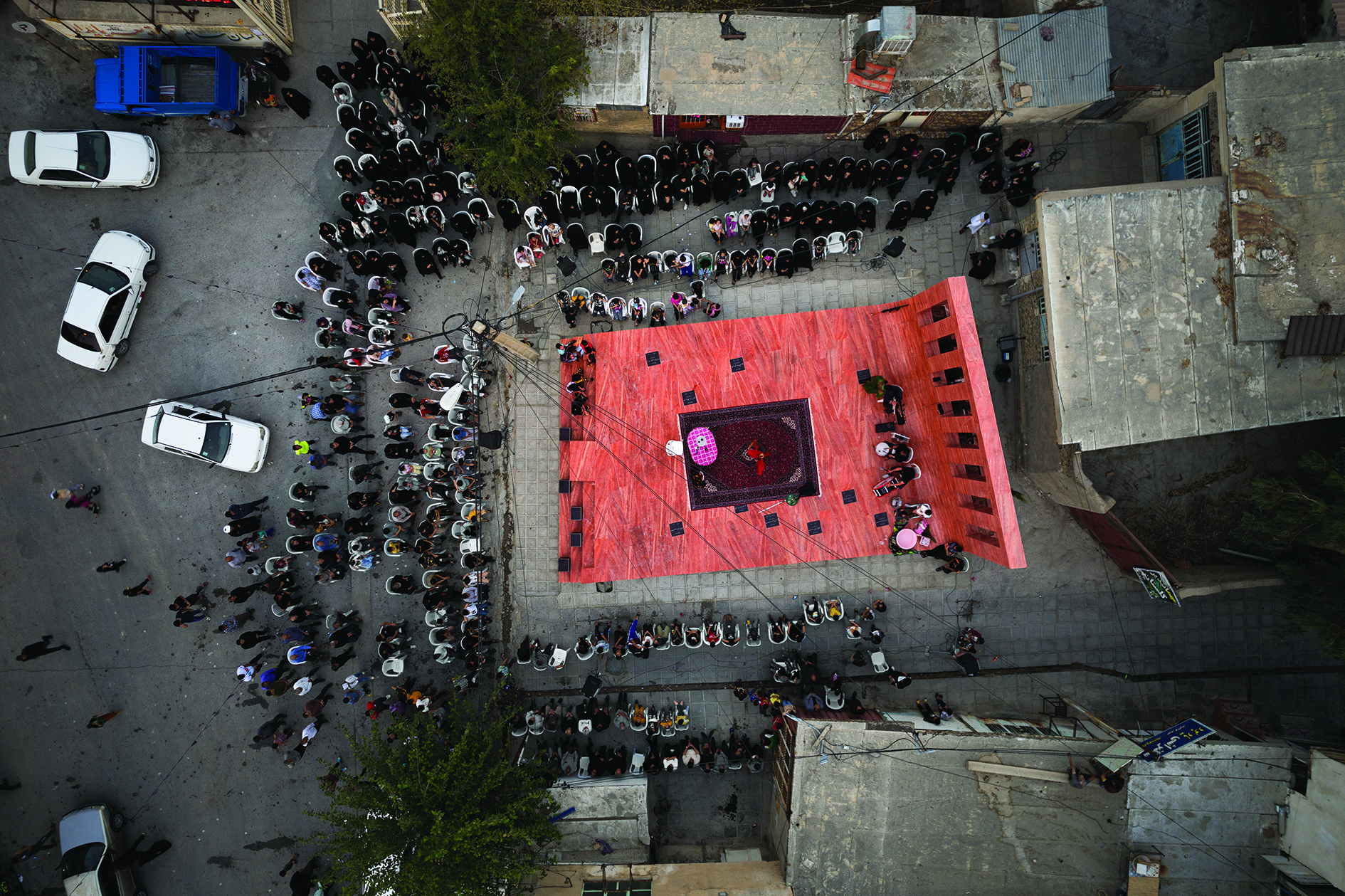 Aerial view of a Taziyeh religious ceremony on the red platform with mourners gathered around, black-clad women on one side, men on the other, and the town's fabric visible around the edges