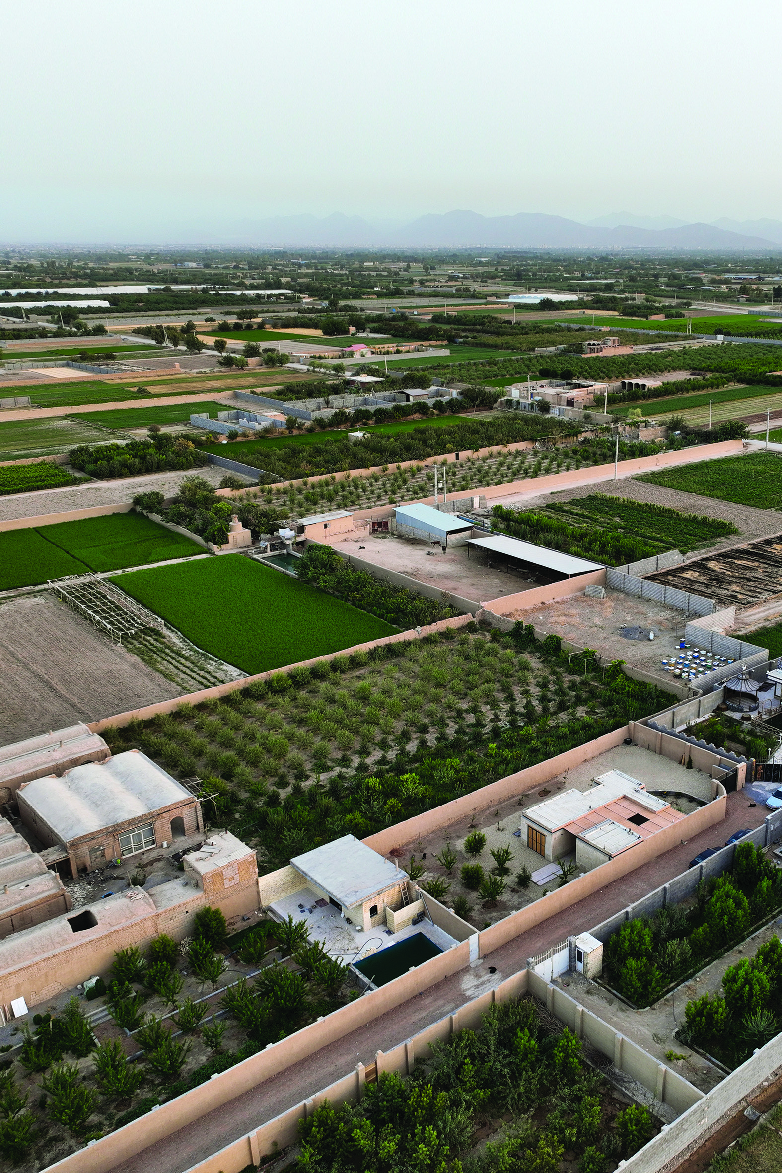 Aerial view of Fahran village showing agricultural fields, orchards, and the house nestled among traditional mud-brick structures with mountains in the distance
