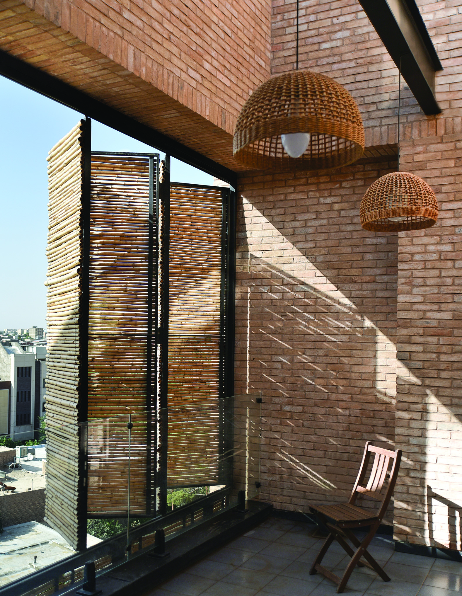 A terrace with open reed panels revealing the city view beyond, wicker pendant lamps hanging from the ceiling, brick walls, and a wooden folding chair