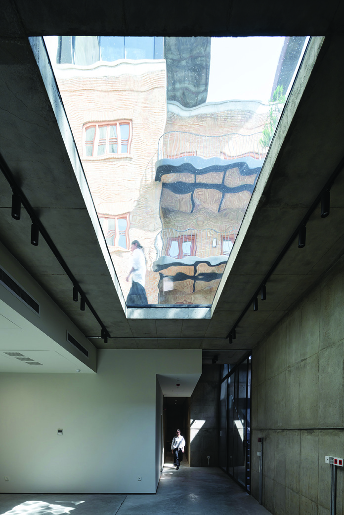 Basement corridor with exposed concrete walls and a glass ceiling panel showing the distorted reflection of the brick facade and courtyard above