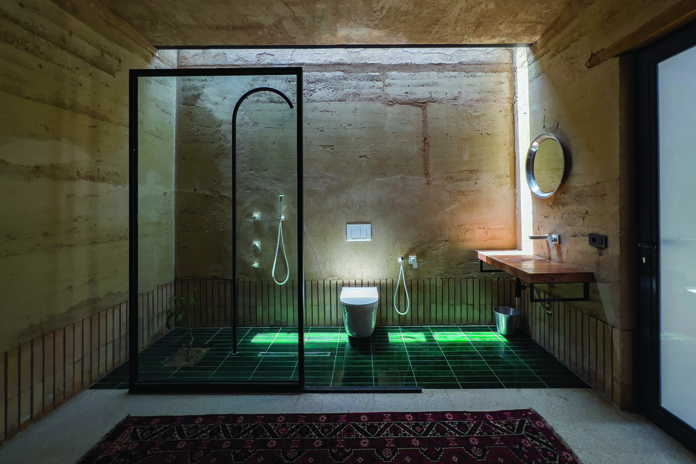 Bathroom with green tile floor, glass shower partition, rammed earth walls, round mirror, and warm underlighting