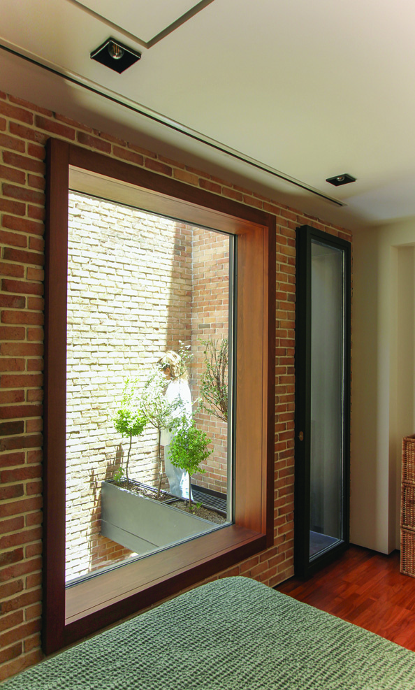 A bedroom with a large wood-framed window looking into the brick lightwell with planted greenery, beside a glass door to adjacent space