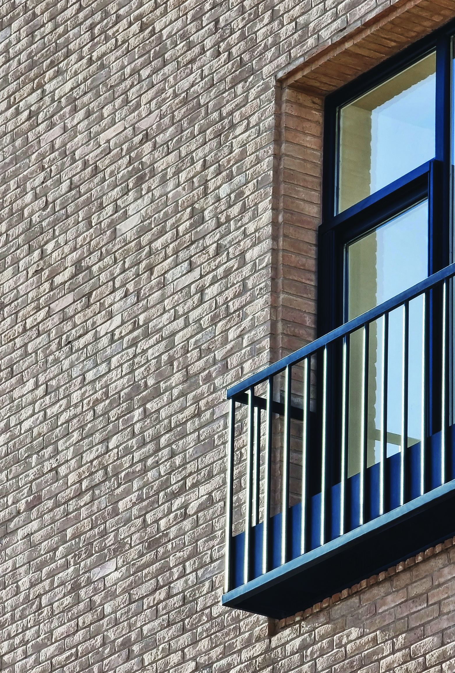 Close-up of the brick facade showing a window with dark metal balcony railing and the texture of locally produced Kerman brick