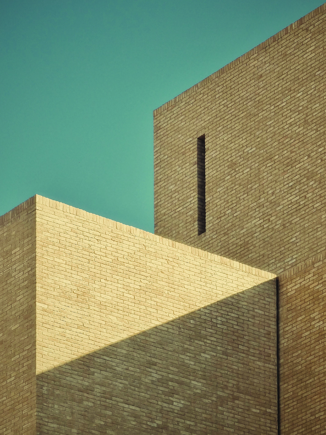 Abstract composition of brick volumes and geometric shadows against a turquoise sky, showing the interplay of mass and void on the upper floors