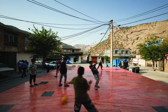 Children playing on the red platform with mountains visible in the background, power lines overhead, and the small-town fabric of Sepiddasht surrounding them