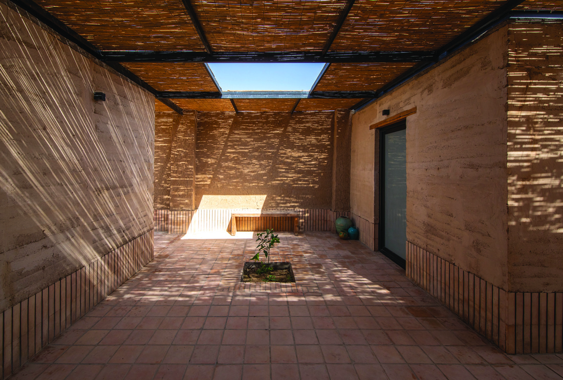Wide view of the central courtyard showing rammed earth walls, reed canopy with skylight, wooden bench, and a young tree