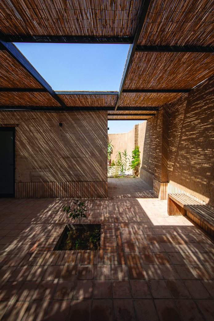 View along the courtyard with a reed canopy overhead, skylight opening, wooden bench, and small trees in planting beds