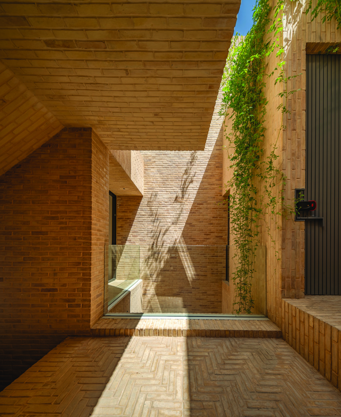View looking up through the brick-clad lightwell showing angular brick volumes, a glass floor panel, climbing greenery, and blue sky above