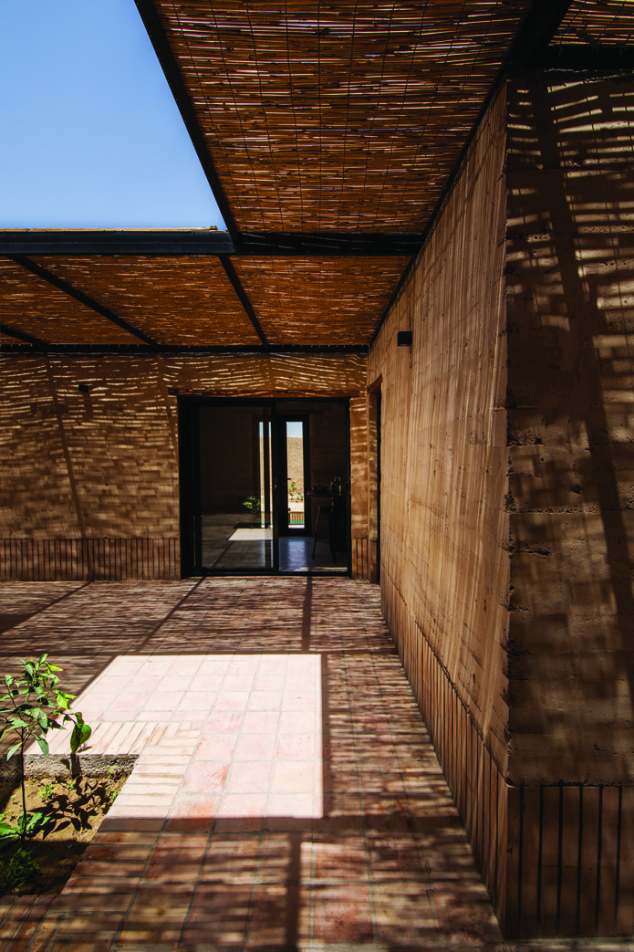 Covered courtyard with intricate reed canopy casting striped shadows on the brick walls and tile floor, with a glass door visible at the end