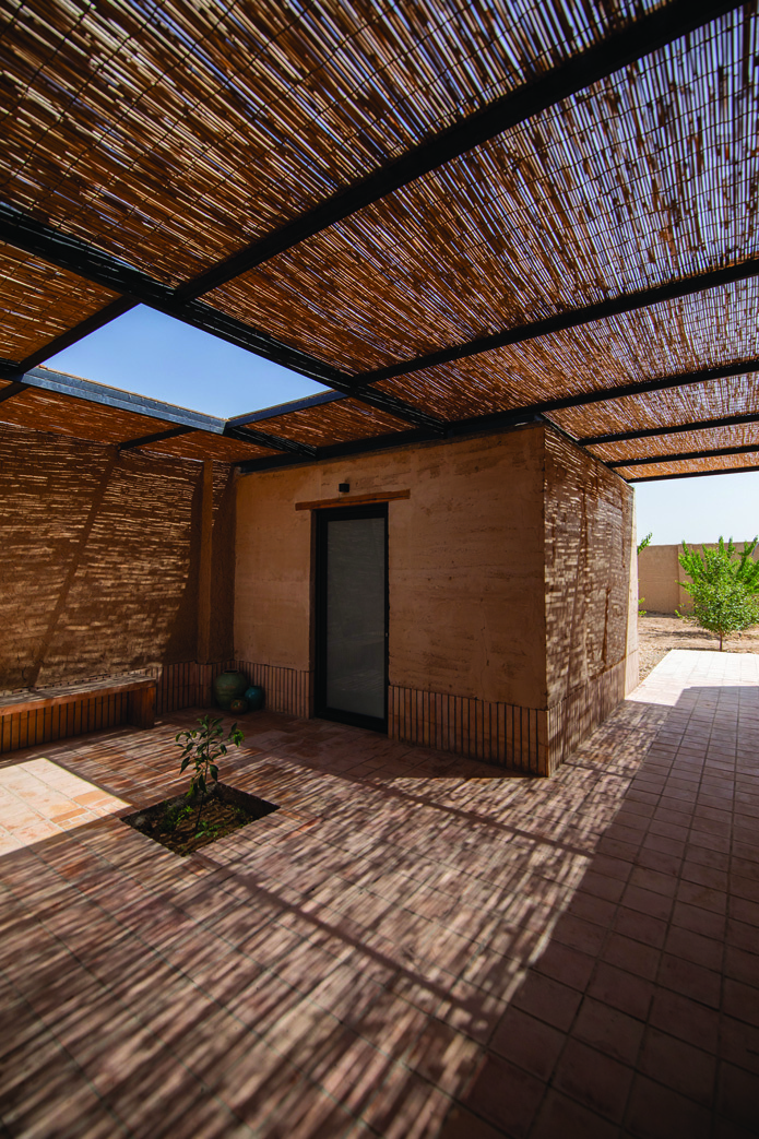 Courtyard view with reed canopy, rammed earth walls, a small tree, and a doorway leading into the interior spaces