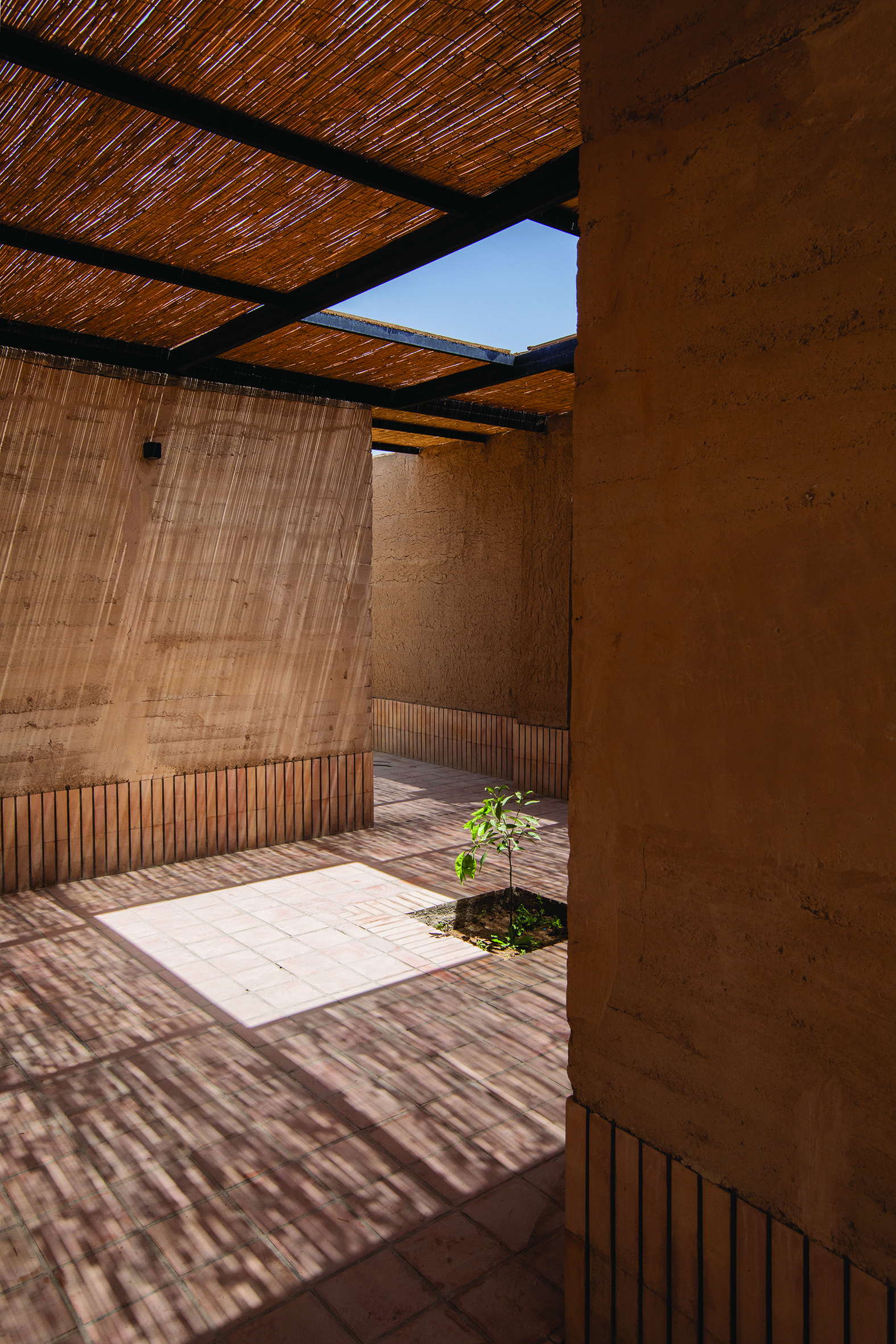 Semi-outdoor courtyard space with reed canopy ceiling, rammed earth walls, and a young tree growing from a planting bed in the tiled floor