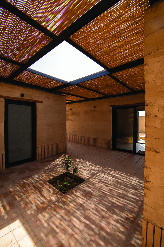 Central courtyard with a skylight opening in the reed canopy, rammed earth walls, and a small tree in a planting bed at the center