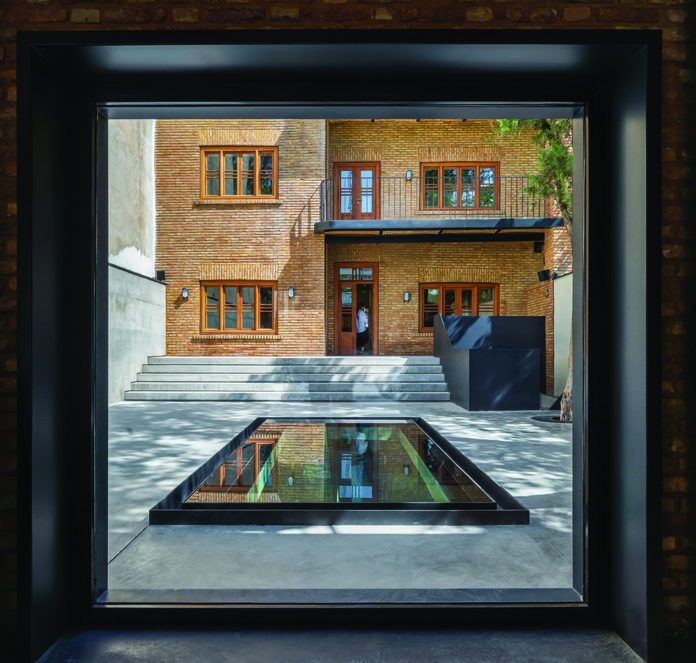 View of the courtyard through a large window frame, showing the brick facade with wooden windows, stone steps, and a glass floor panel reflecting the building above